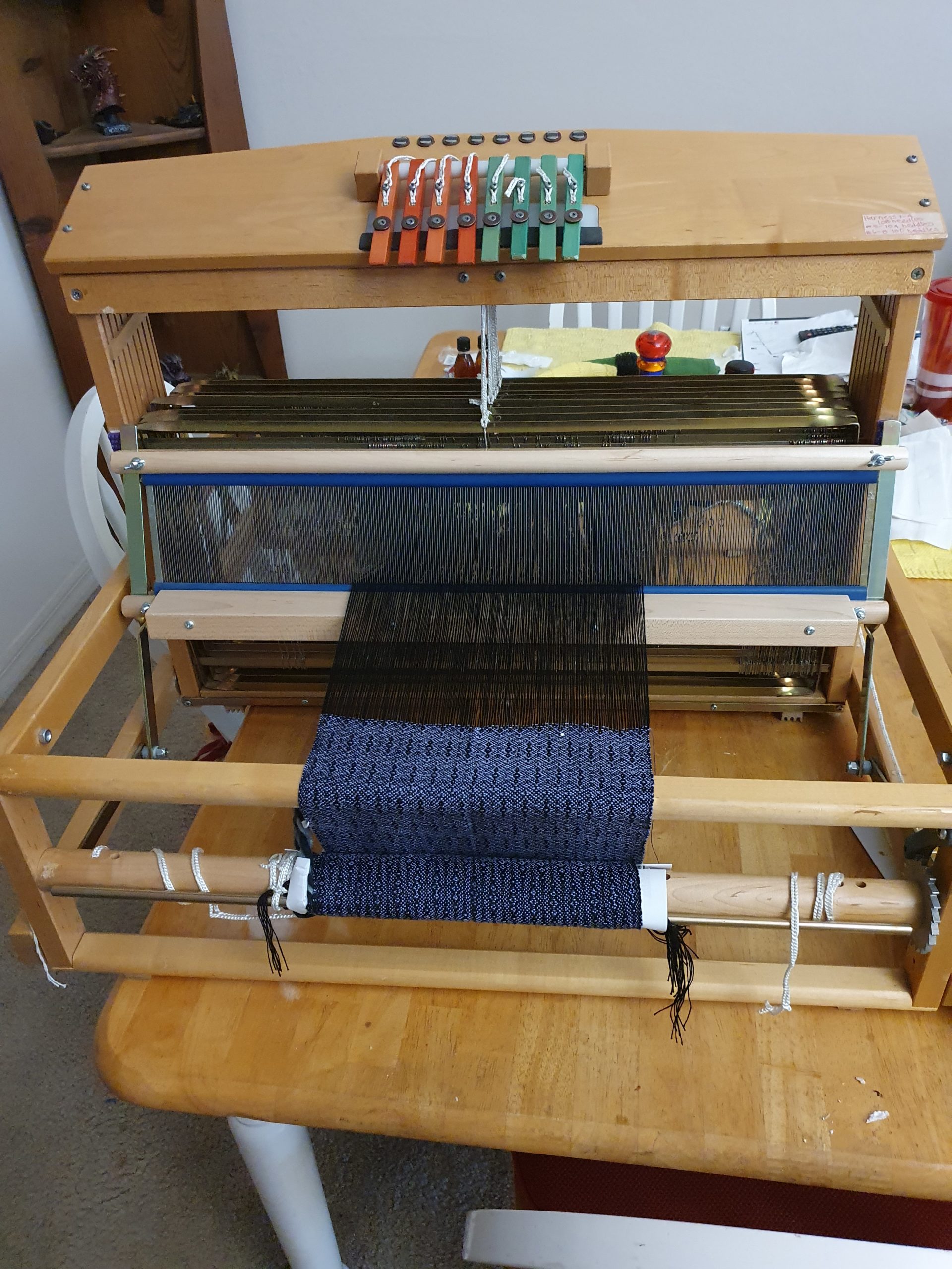 A weaving loom sitting on a dining table. There's a purple and black scarf project on it.